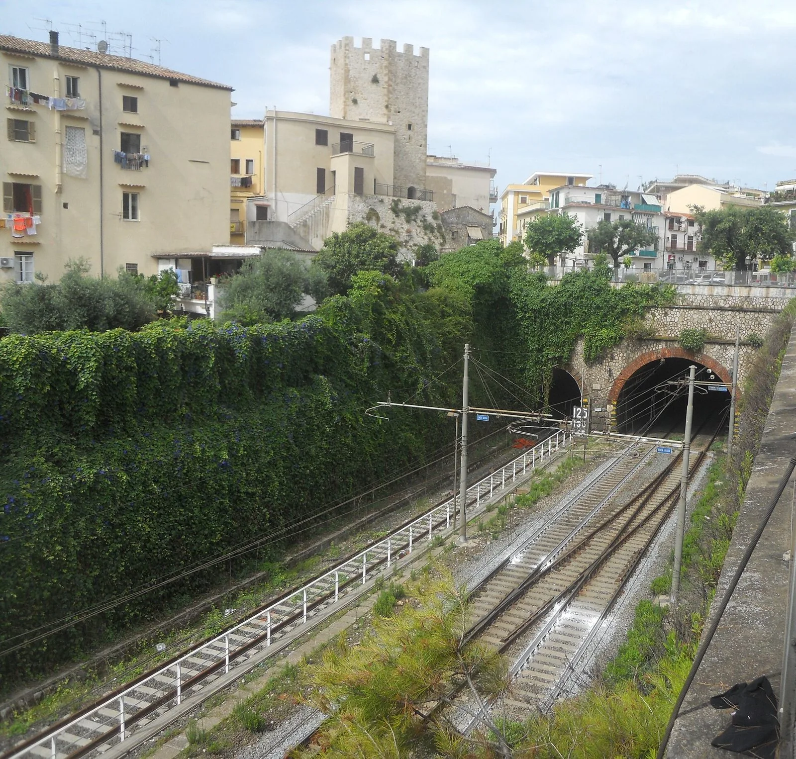 I binari della stazione di Formia-Gaeta in trincea tra la vegetazione, con le gallerie e una torre medievale sullo sfondo